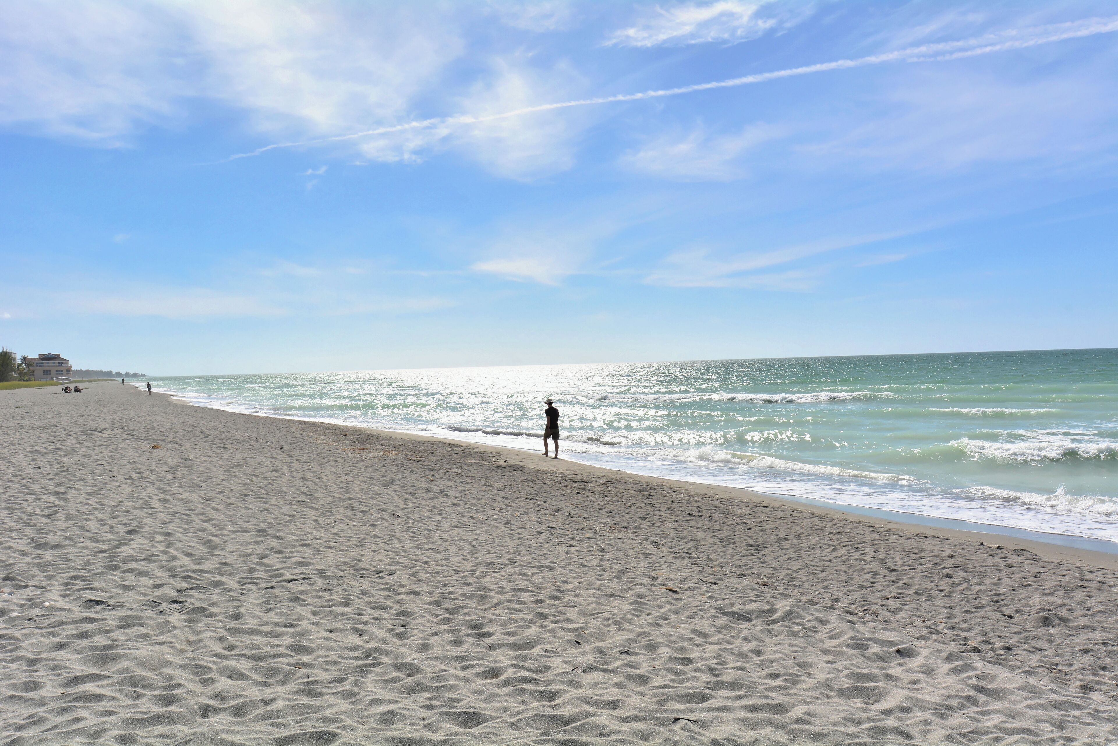 Ein Strand mit einer Person, die am Ufer entlanggeht, wolkenlosem Himmel und ruhigen Wellen.