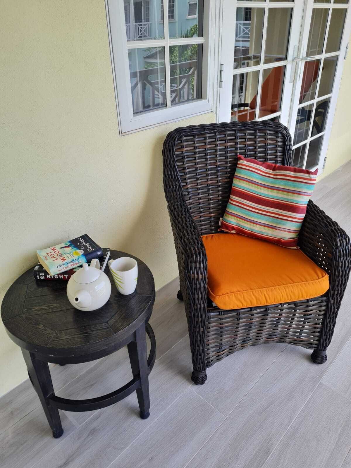 Ein Korbsessel mit orangefarbenem Kissen und einem gestreiften Kopfkissen, ein kleiner runder Tisch mit Teekanne und Büchern sowie ein Fenster mit Blick auf Balkone.