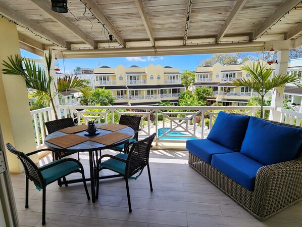 A covered outdoor seating area with a table and chairs, a blue sofa, and a view of a pool and buildings in the background.