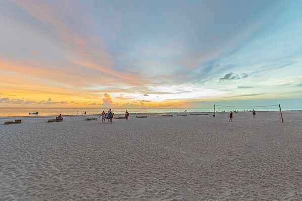 Ein Beachvolleyballfeld mit Spielern und Zuschauern, umgeben von Sand und einem Sonnenuntergangshimmel.