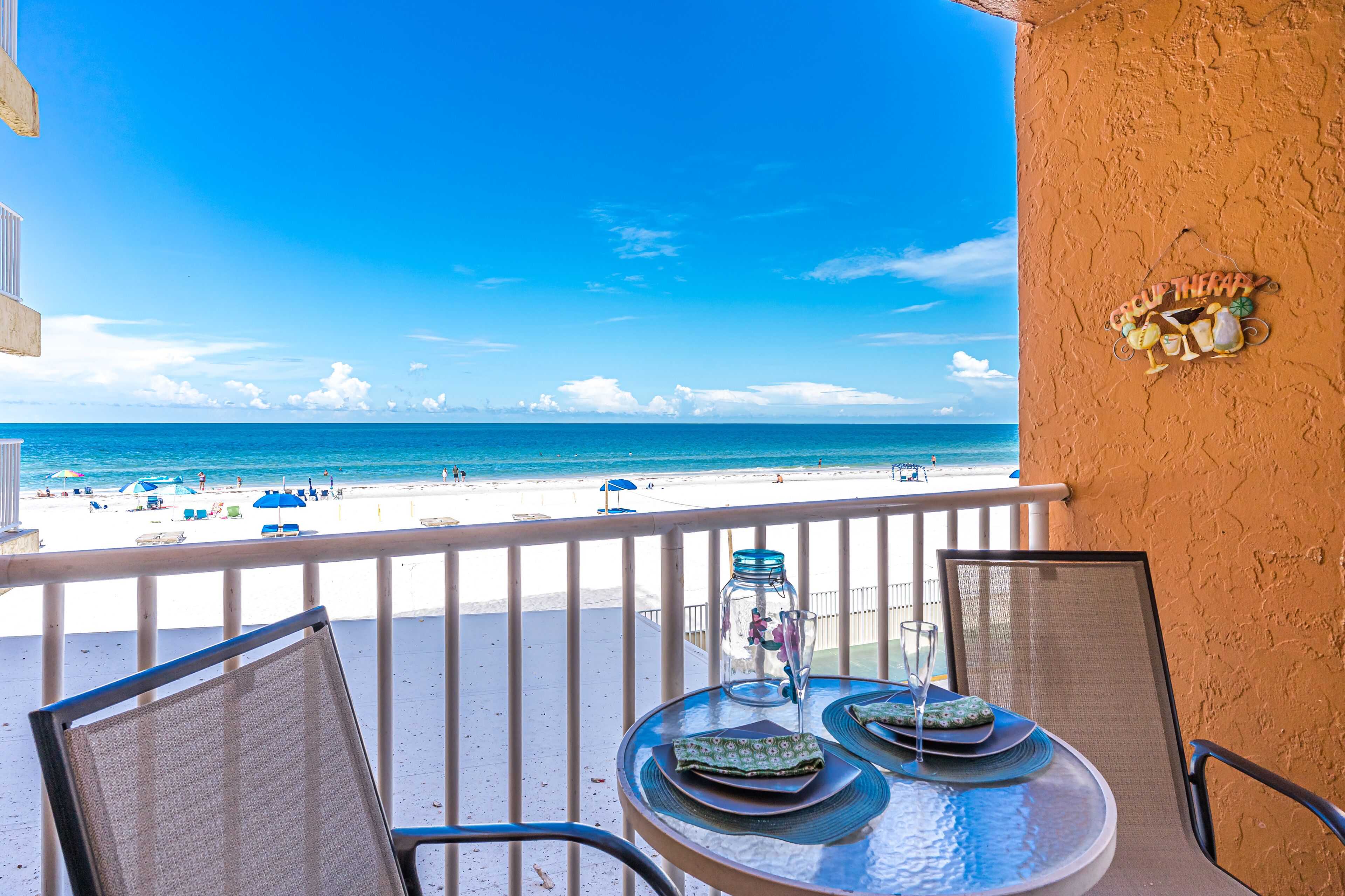 A balcony with a table set for two, overlooking a beach with umbrellas and people.
