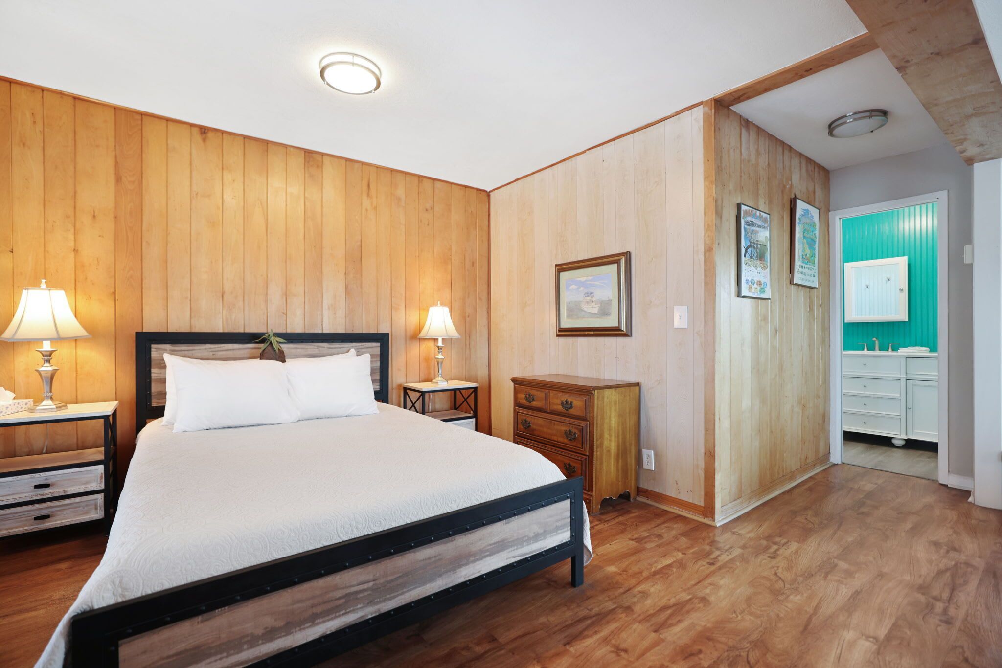 Ein Schlafzimmer mit einem Bett mit Holz-Kopfteil, zwei Nachttischen mit Lampen, einer Kommode und einem Badezimmer mit grüner Wand und weißen Armaturen.