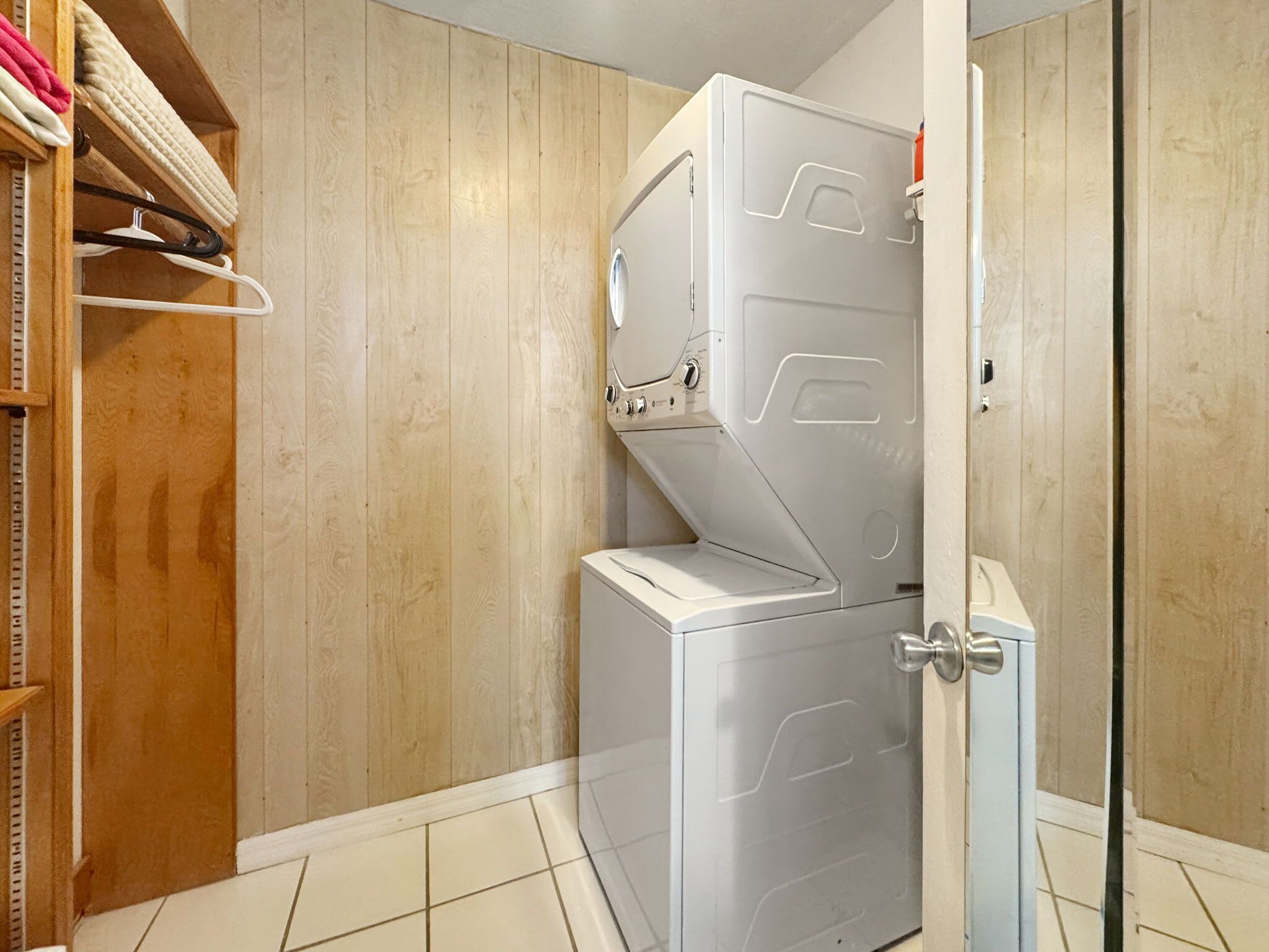 Une buanderie équipée d’un lave-linge à chargement frontal, d’une armoire en bois où sont suspendus des vêtements, et d’un sol carrelé.
