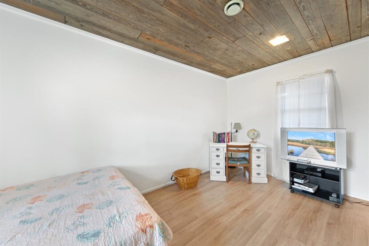 Una camera da letto con soffitto in legno, un letto con copripiumino floreale, una piccola scrivania con libri, una televisione e una finestra con persiane.