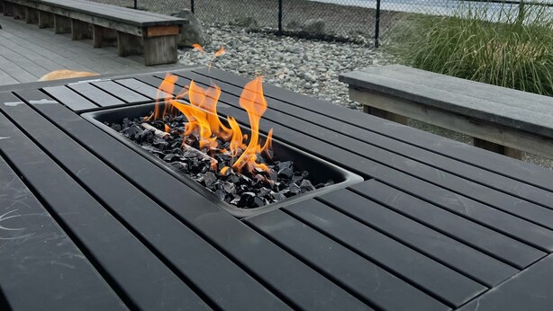 A fire pit with flames, surrounded by a wooden deck with benches, overlooking a body of water and a rocky shoreline.