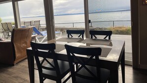 A dining area with a view of the sea, featuring a table with four chairs, a brown sofa, and a glass partition.