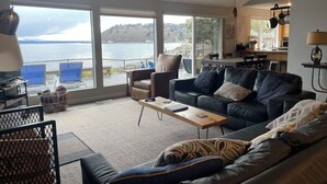A living room with a large window overlooking a body of water, a black leather sofa, a coffee table, and a kitchen area visible through the window.