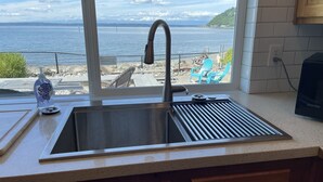 A kitchen with a stainless steel sink, a window overlooking a beach, and a bottle of hand sanitizer on the counter.