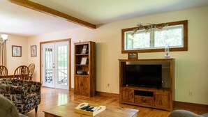 A living room with a wooden entertainment center, a sofa, a coffee table with remote controls, and a large window with a view of greenery. - Pet Friendly Red House: Short Walk to Private Dock & Lake George!  (Lake George)