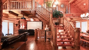 Interior de una cabaña de troncos con una escalera de madera, un comedor y una sala de estar con sofá y sillones.