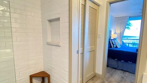 Ein Badezimmer mit Dusche, einem hölzernen Hocker und einer Fliesenwand. Eine Tür führt zu einem Schlafzimmer mit Bett und Vorhängen.
