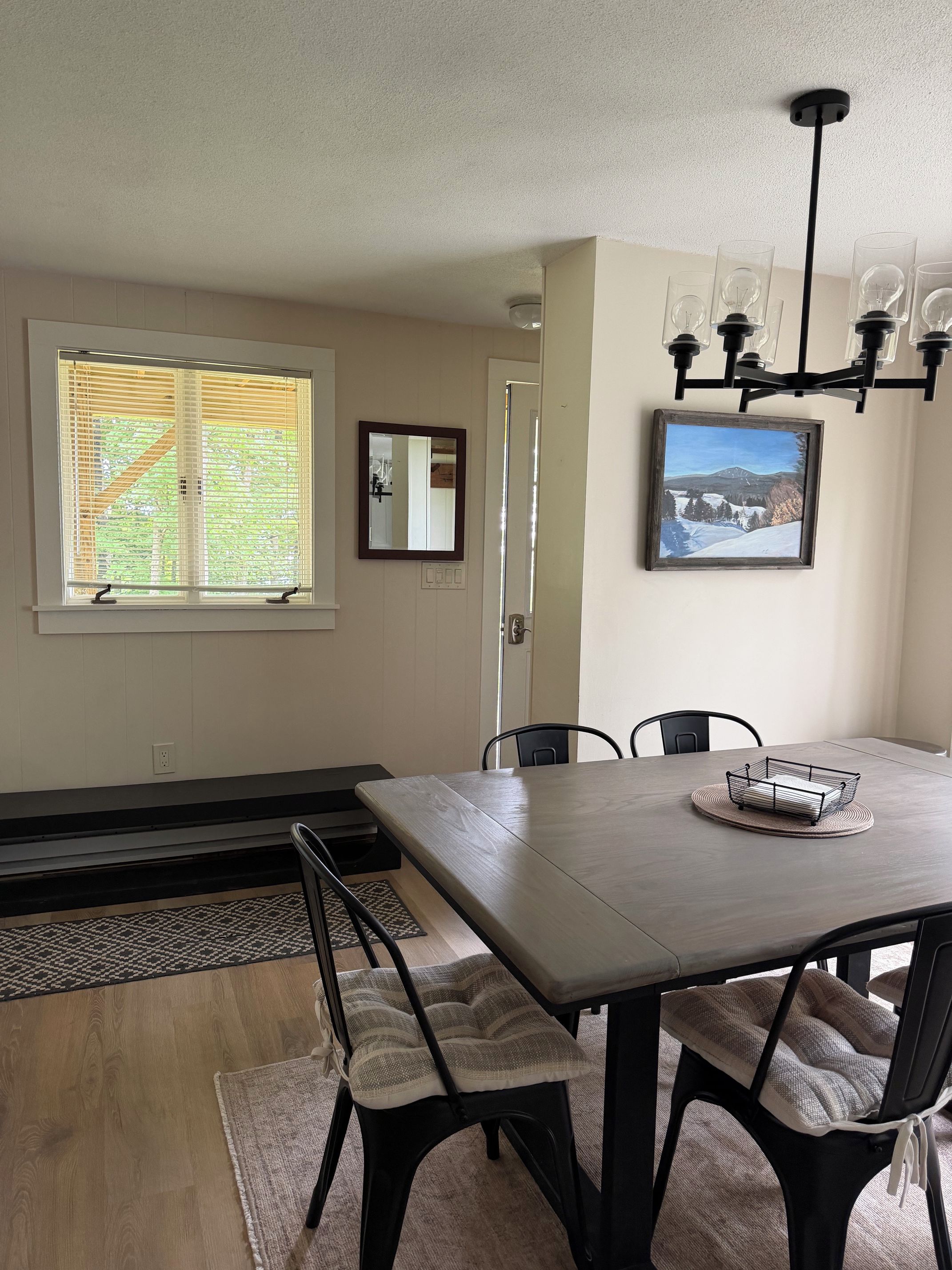 A dining area with a table, chairs, a chandelier, a window with blinds, a mirror, and a framed picture on the wall.