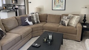 A living room with a sectional sofa, a coffee table, and a kitchen in the background. - Ski On, Ski off Okemo Trailside Appartment, Sleeps 6/ $550per night, (Ludlow)