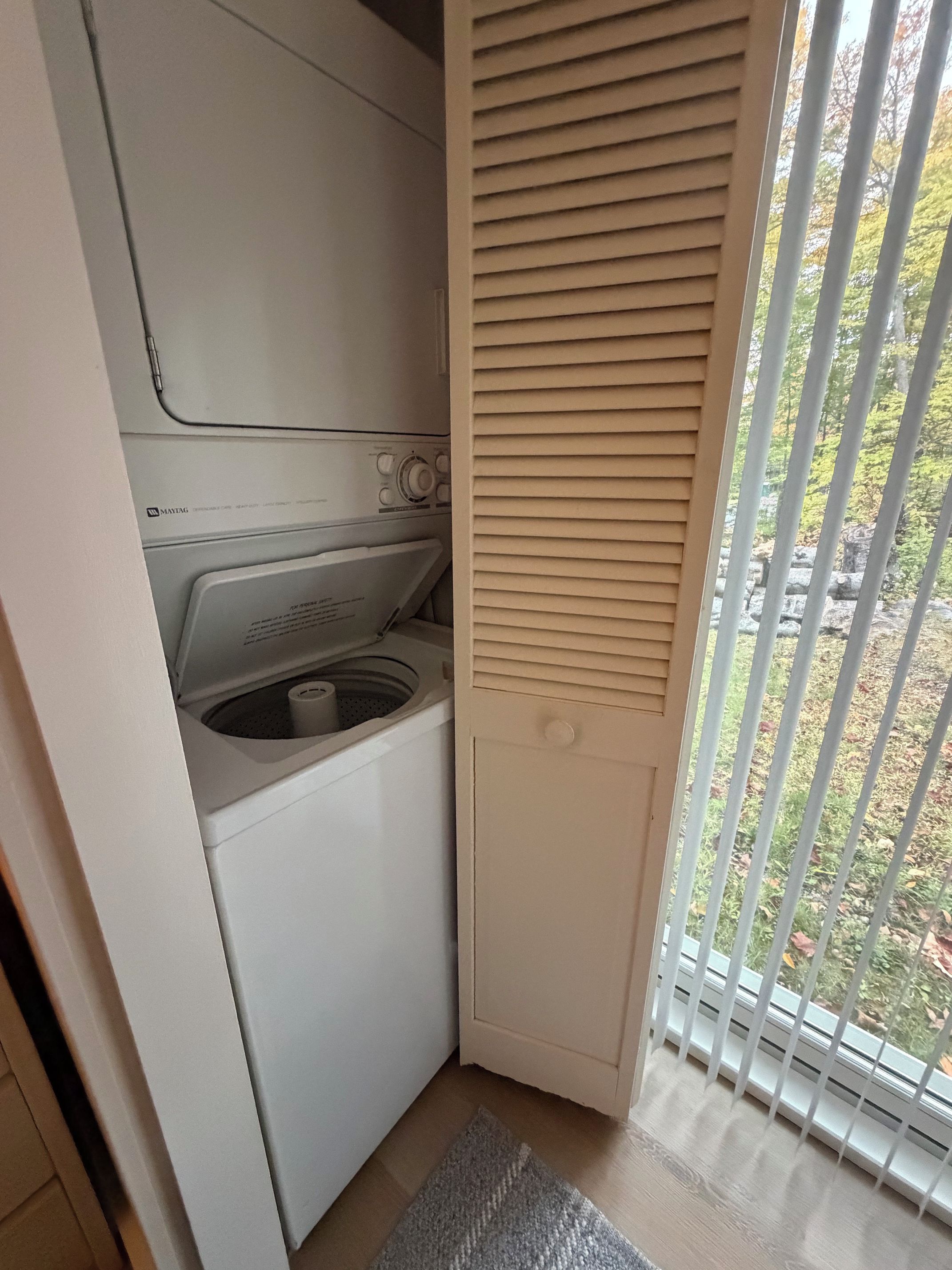 Un espace buanderie compact comprenant un lave-linge et un sèche-linge, une petite fenêtre avec des stores et une vue sur les arbres à l’extérieur.