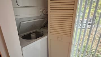 A compact laundry area with a washing machine and dryer, a small window with blinds, and a view of trees outside.