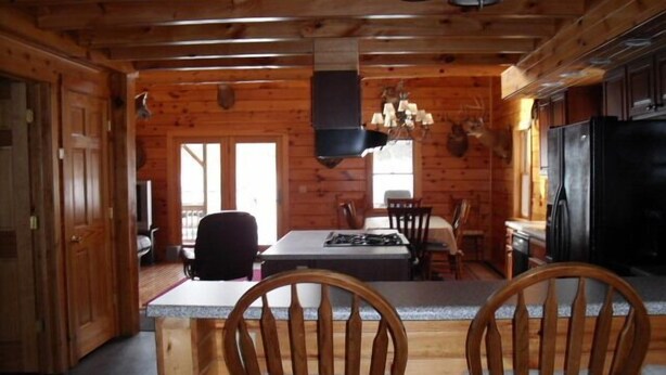 A wooden cabin interior with a dining table, chairs, a kitchen area, and a large window. - Log Cabin-Unique/Beautiful (Candor)