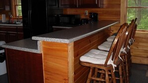 A kitchen with wooden cabinets, a black refrigerator, a granite countertop, and a bar area with wooden stools. - Log Cabin-Unique/Beautiful (Candor)