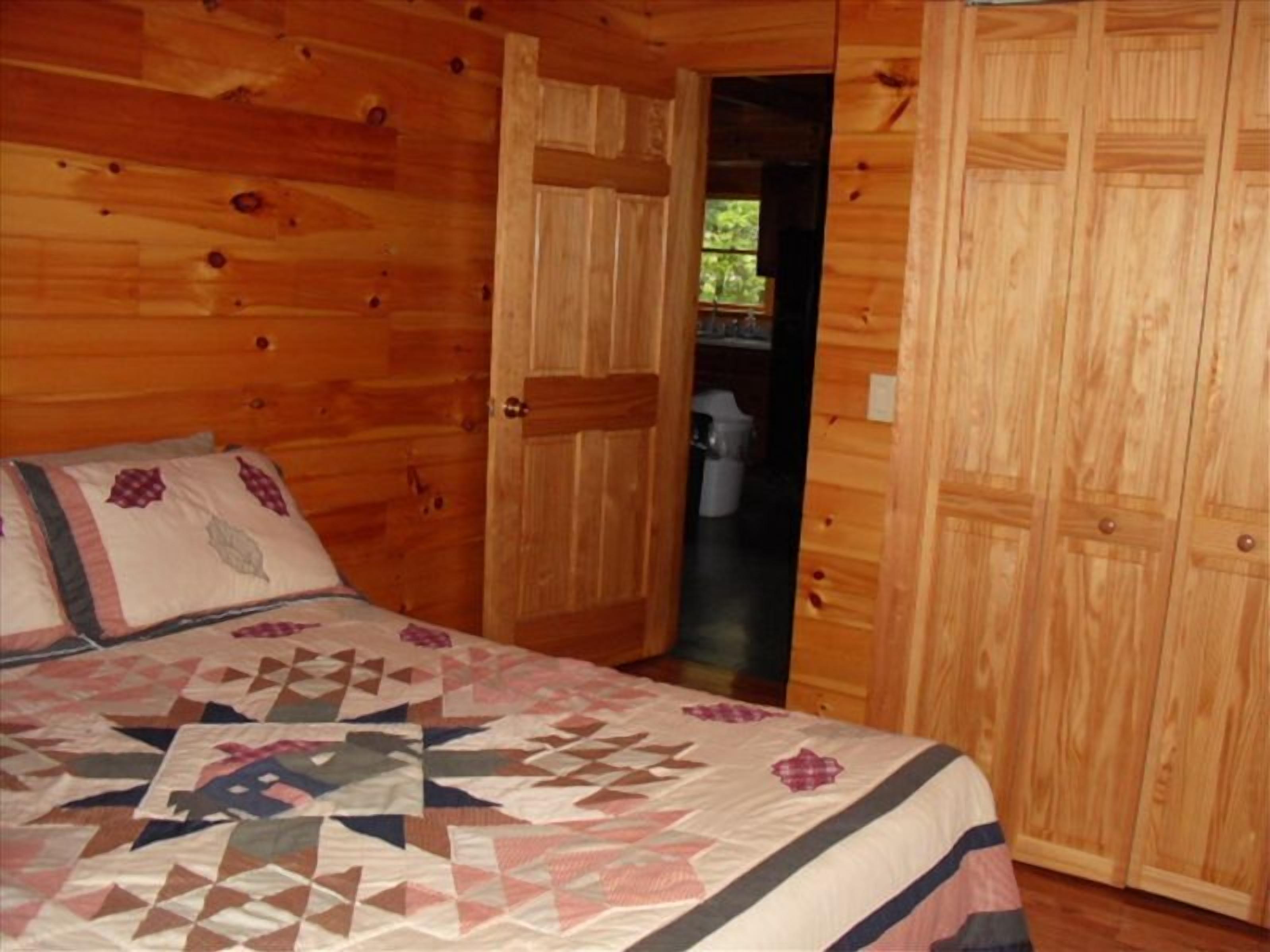 A wooden cabin bedroom with a bed, a quilt, and a door leading to another room.