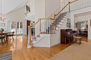 A spacious living area with a wooden staircase, a piano, and a dining area with chairs and a table. - Chatham-Walk to Town/Beach-Multi Family Friendly (Chatham)