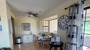 Un vaste espace dédié à la salle à manger, comprenant une table et des chaises, un ventilateur de plafond, une horloge et une vue sur l’extérieur à travers une fenêtre munie de stores.