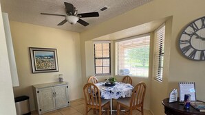 Un coin repas comprenant une table ronde, des chaises, un ventilateur de plafond, une horloge et une fenêtre avec des stores.