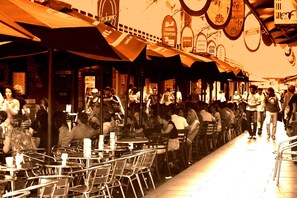 Un café al aire libre animado con mesas y sillas de metal, rodeado de sombrillas y señales que indican una zona céntrica y espacios para leer.