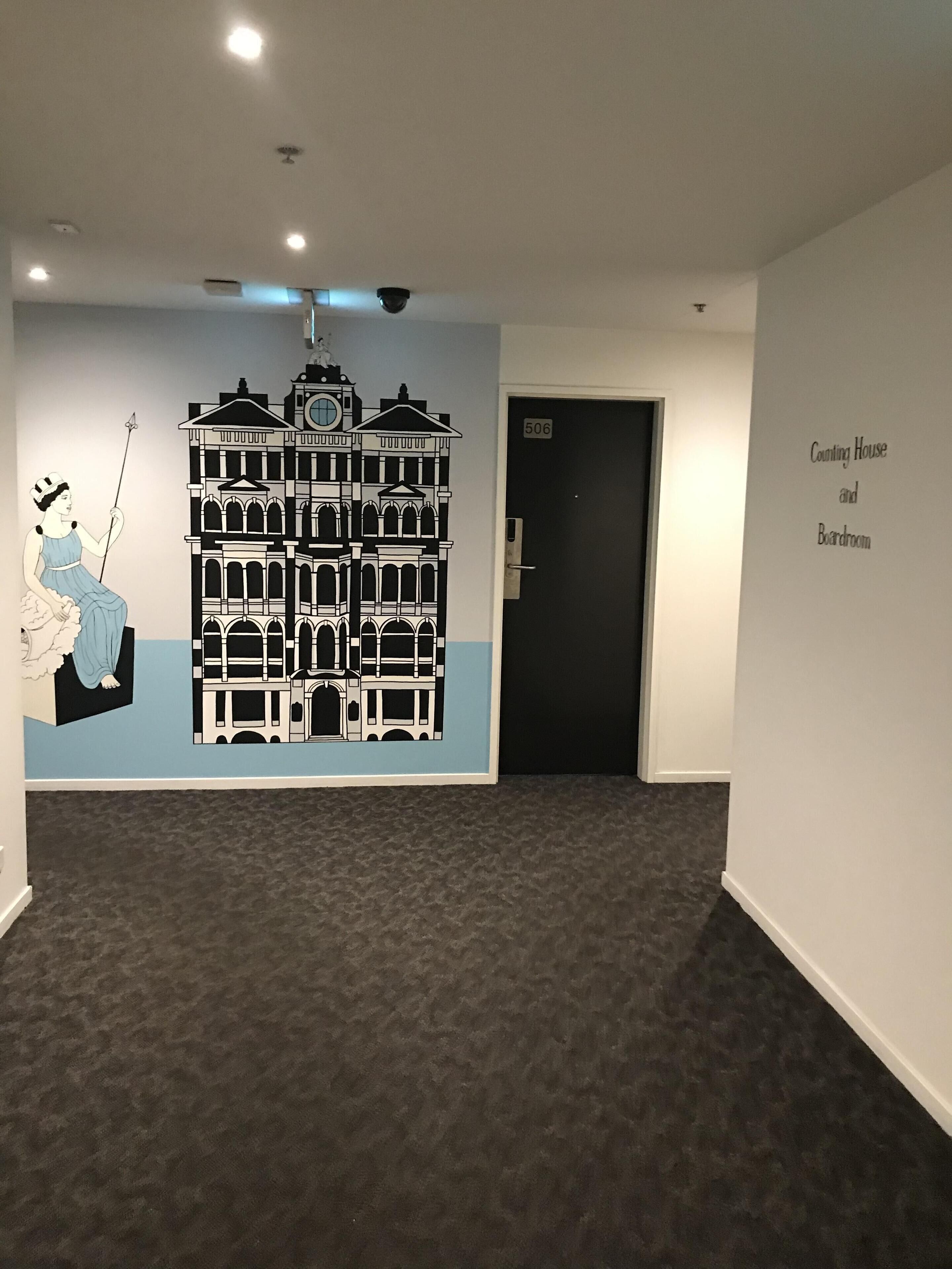 Een gang met een zwart-wit muurschildering van een gebouw en een vrouw op een schommel, een deur met daarop '506' en een bordje met de tekst 'Counting House and Boardroom'.