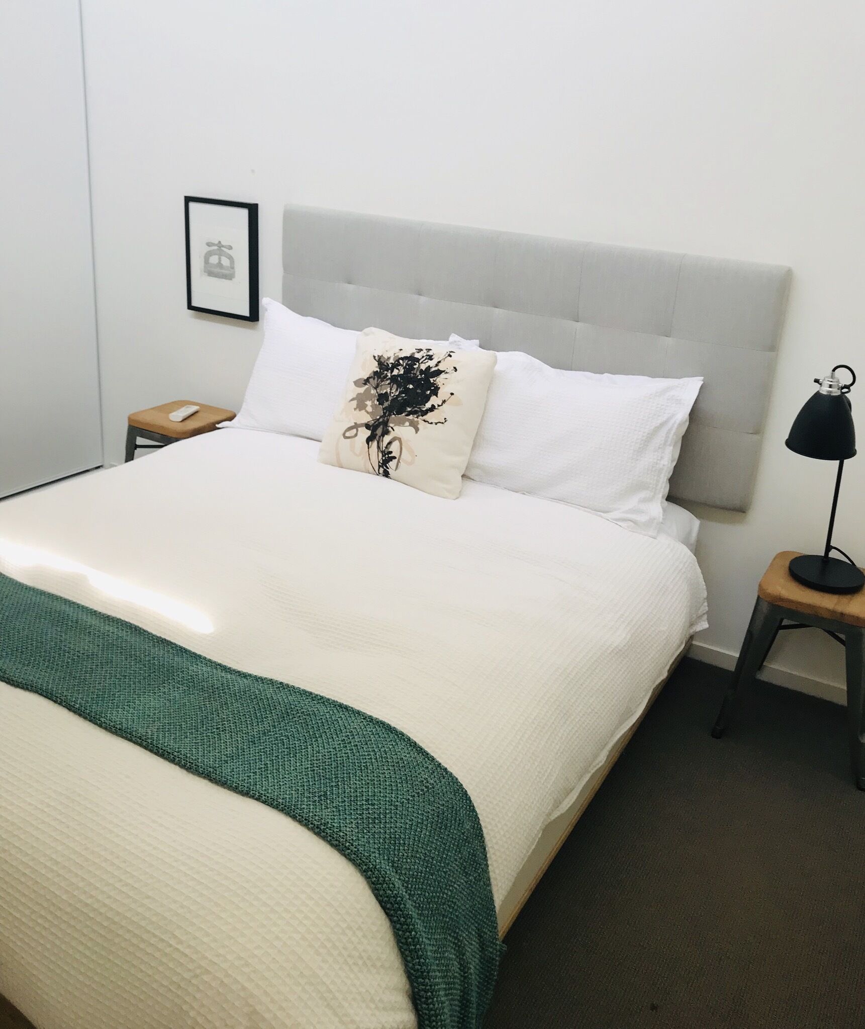 A neatly made bed with a white headboard, a green throw blanket, and a decorative pillow with a black floral design. There is a bedside table with a lamp and a small wooden stool.