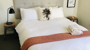 Un llit ben fet amb llençols blancs, una manta marró i un coixí decoratiu. Hi ha una petita taula de nit de fusta amb una làmpada i un comandament al costat del llit.