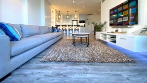 Una sala moderna con un sofá gris, una mesa de centro y una alfombra. Se ve una cocina al fondo con un comedor.