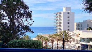 Un hotel frente al mar con vistas al océano, presenta un edificio moderno de varios pisos y balcones, rodeado de palmeras y vegetación.