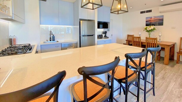 Una cocina moderna con una isla central, electrodomésticos de acero inoxidable, iluminación colgante y un comedor con sillas de madera y una mesa.
