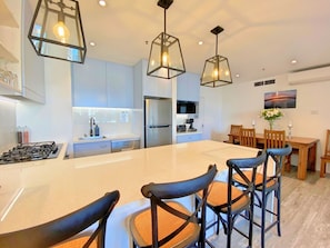 Una cucina moderna con isola centrale, elettrodomestici in acciaio inossidabile, lampade a sospensione e zona pranzo con sedie e tavolo in legno.