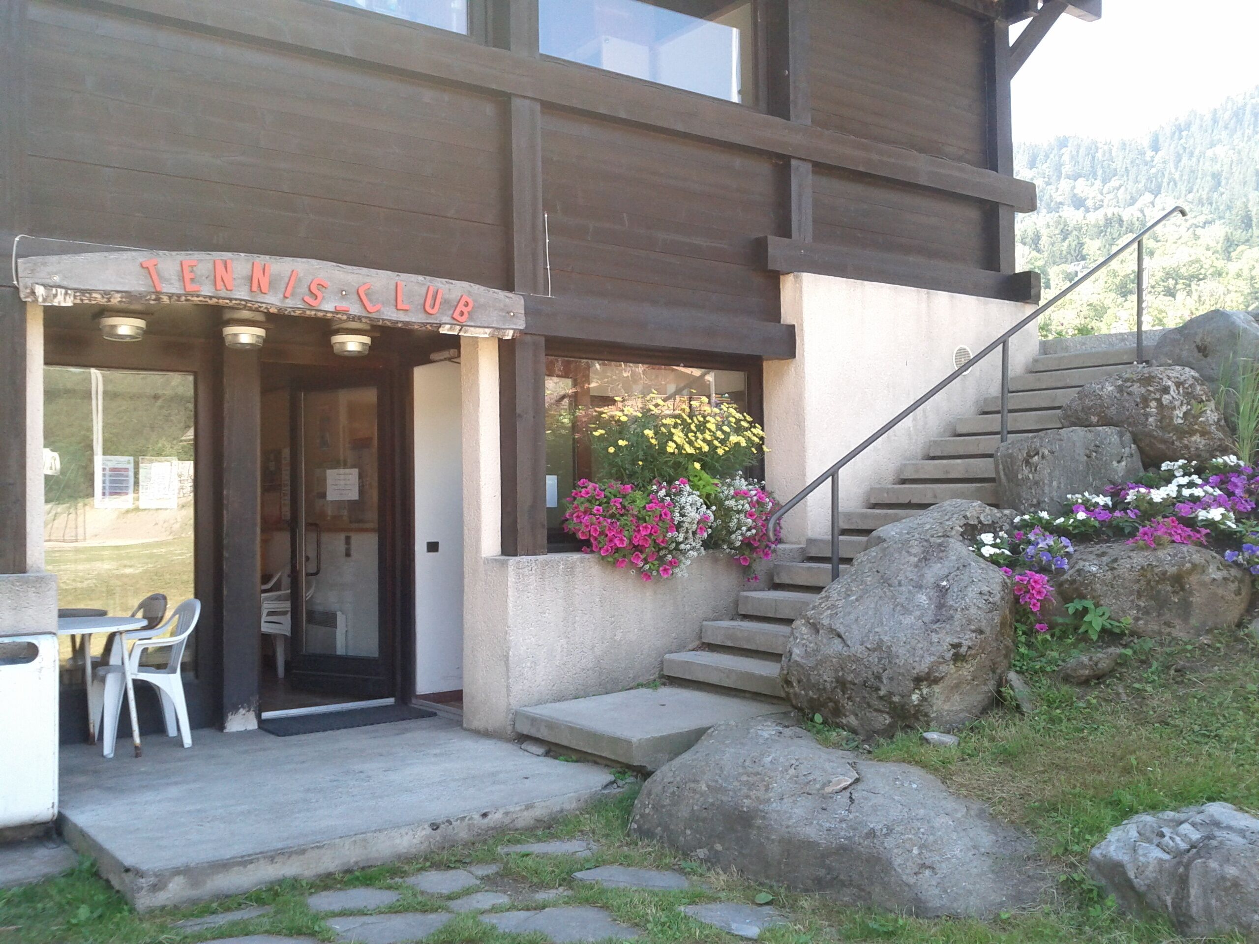 A building with a sign reading "TENNIS CLUB" at the entrance, a staircase with large rocks, and a small outdoor seating area with chairs.