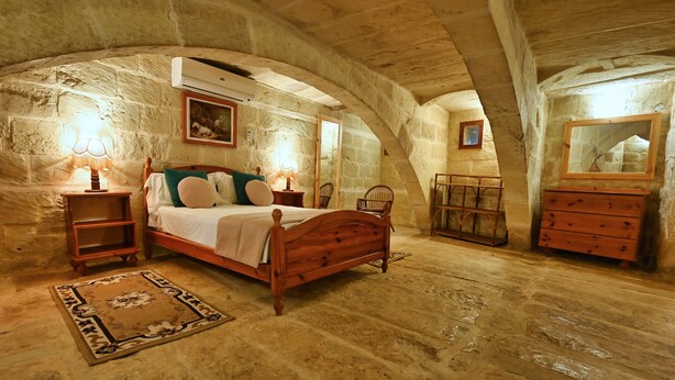A bedroom with a wooden bed, two bedside tables with lamps, a dresser, and a mirror. The room has stone walls and an arched ceiling.