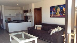 A living room with a dark brown sofa, a glass coffee table, and a kitchen area in the background. - Apartment with Large Sunny Balcony with stunning views (Adeje)