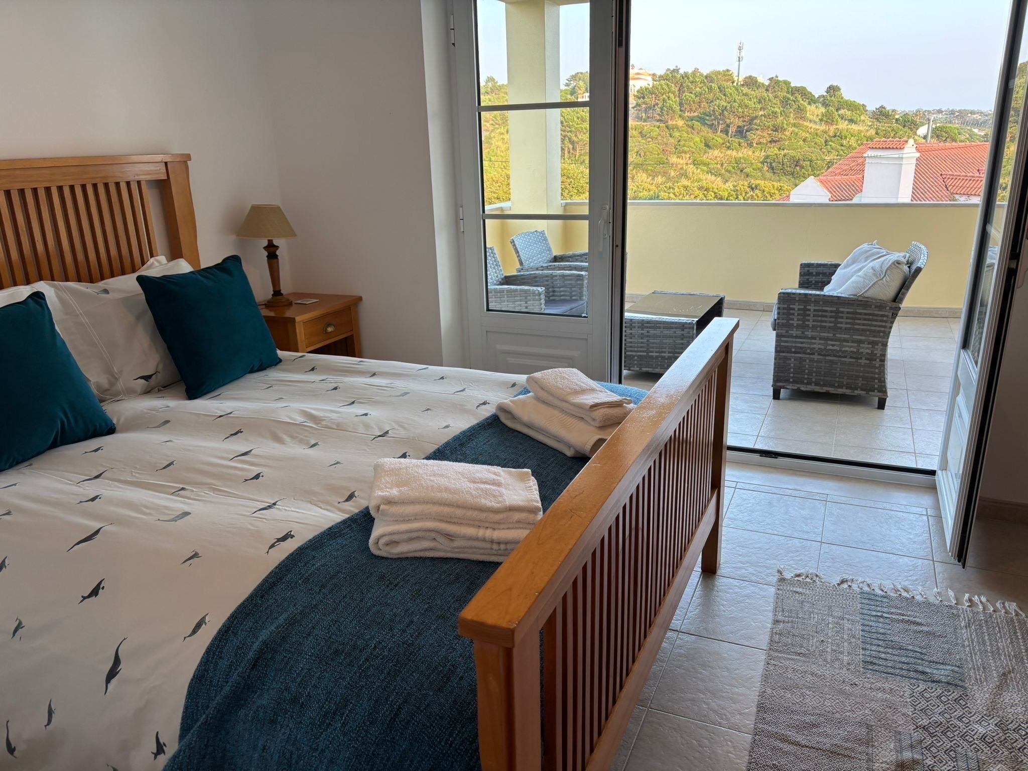 A bedroom with a bed, pillows, a nightstand, a lamp, and a view of a balcony with wicker furniture.
