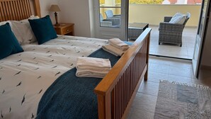 Un dormitorio con cama, almohadas, una mesita de noche, una lámpara y vistas a un balcón con mobiliario de mimbre.