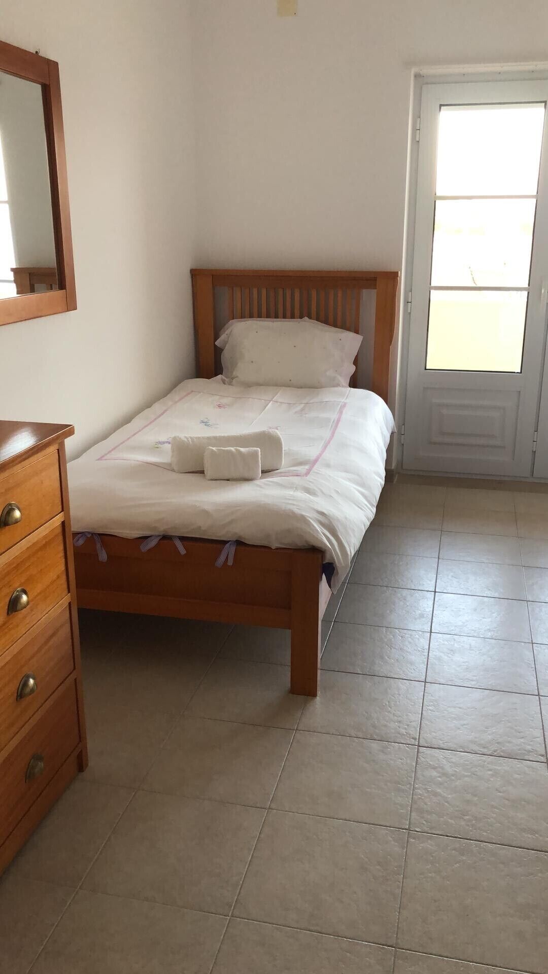 Habitació individual amb un llit i capçalera de fusta, una tauleta de nit amb calaixos i una porta amb finestre.