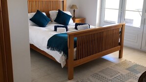 Un dormitorio con cama de madera, ropa de cama blanca, almohadas azules, una mesita de noche con lámpara y un ventanal grande.