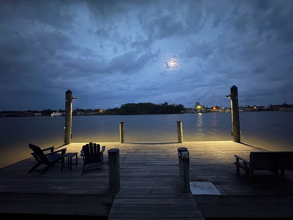 A wooden dock with chairs and a bench, leading to calm water with a moonlit sky.