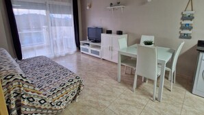 Ein Zimmer mit einem Bett, einem Fernseher, einem Esstisch mit Stühlen, einer kleinen Kommode und einem an der Wand befestigten Regal mit Pflanzen.