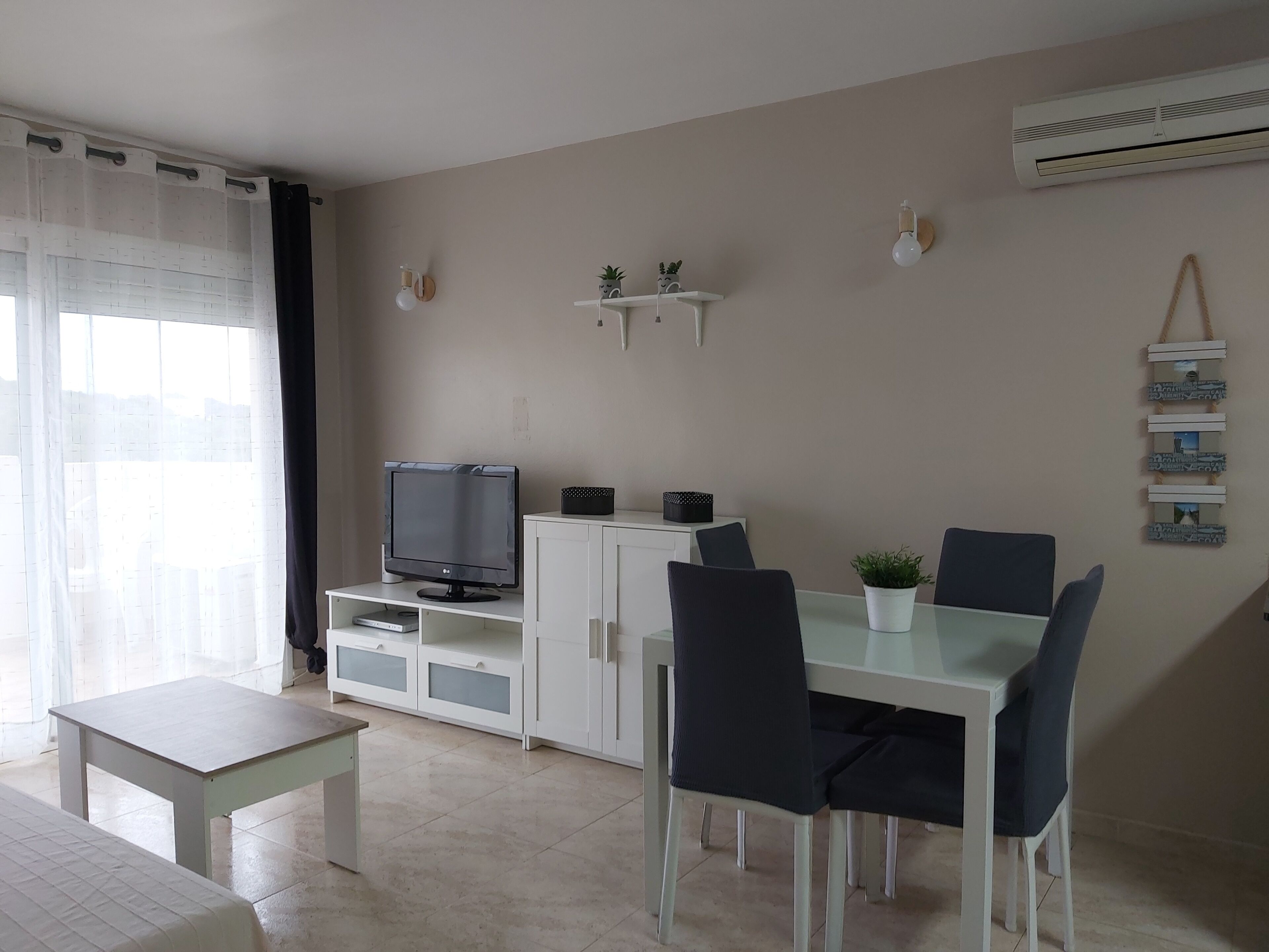 Una sala de estar moderna con un televisor de pantalla plana, armarios blancos, una mesa de comedor de vidrio y sillas oscuras.