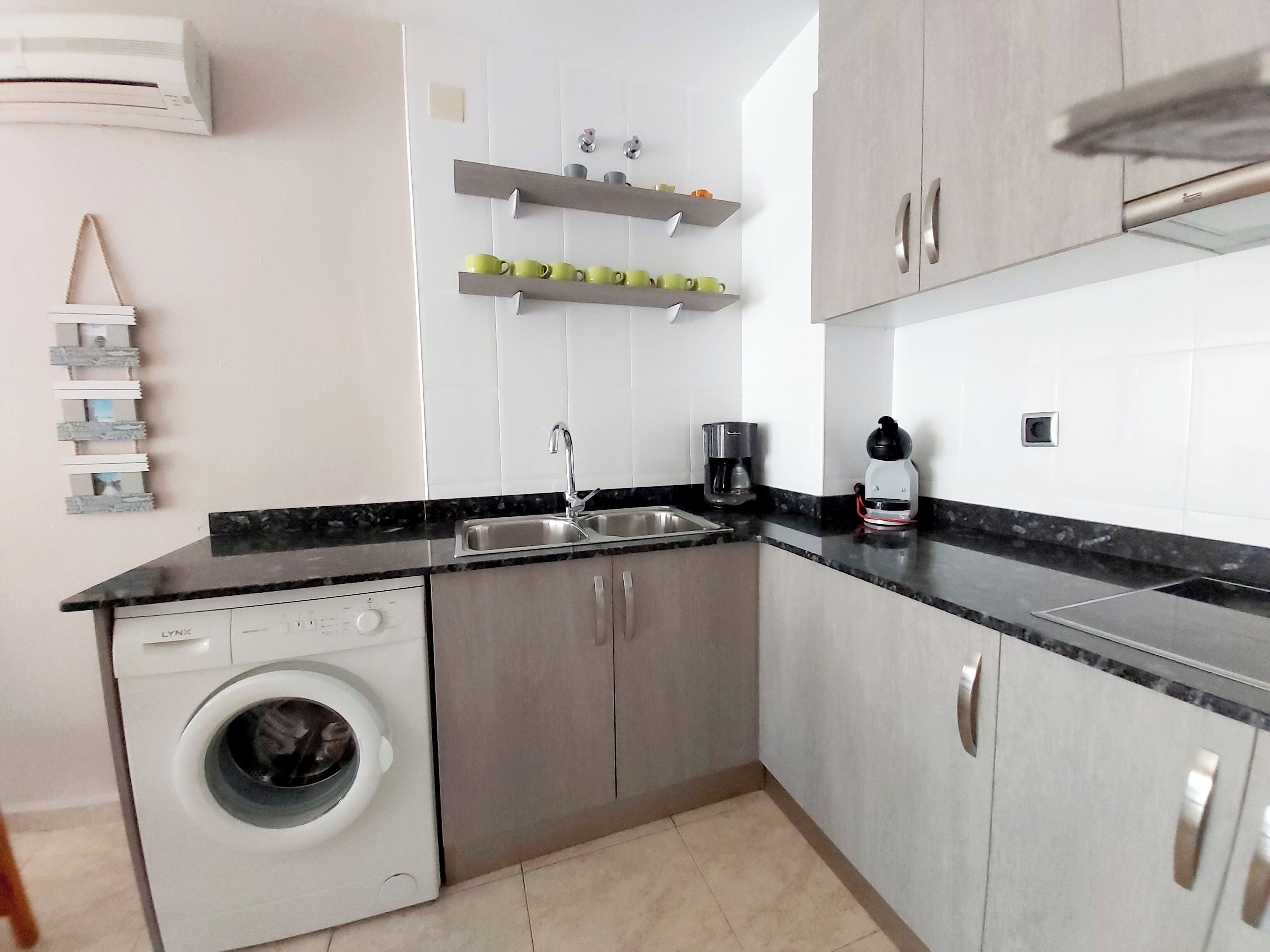 A compact kitchen with a washing machine, a coffee maker, and a shelf with cups.