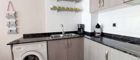 A compact kitchen with a washing machine, a coffee maker, and a shelf with cups.