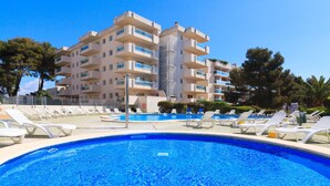 Ein Swimmingpool mit Liegestühlen, ein Gebäude mit mehreren Balkonen und Bäume im Hintergrund.