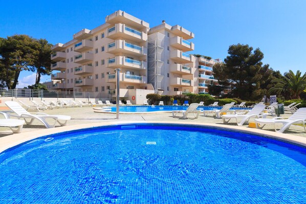 A swimming pool with lounge chairs, a building with multiple balconies, and trees in the background.