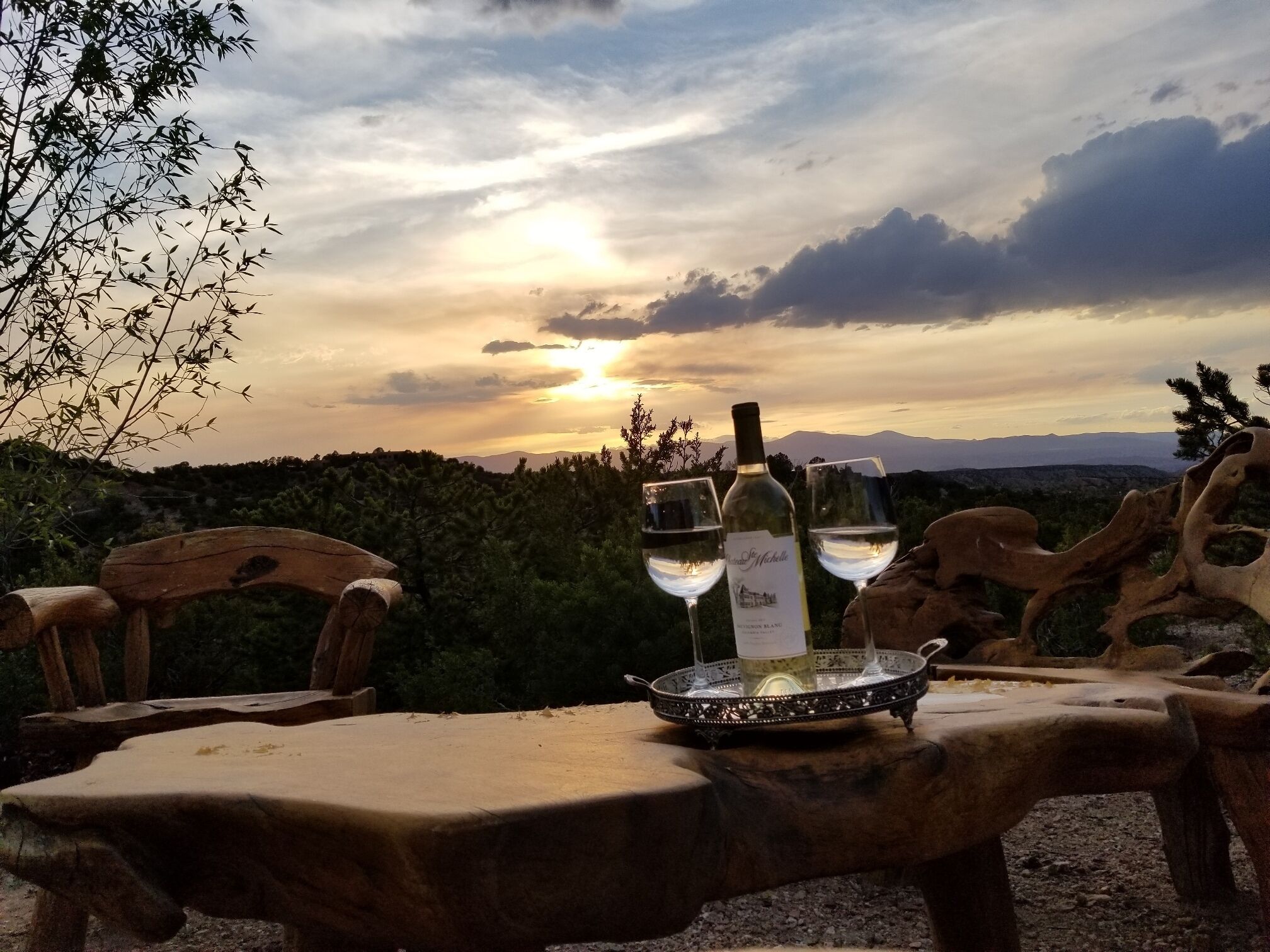 A table with two wine glasses and bottles, set against a scenic sunset view.