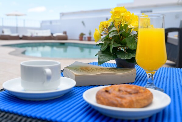 A breakfast setting with a cup, book, flowers, glass of orange juice, and a plate with a pastry by a pool.