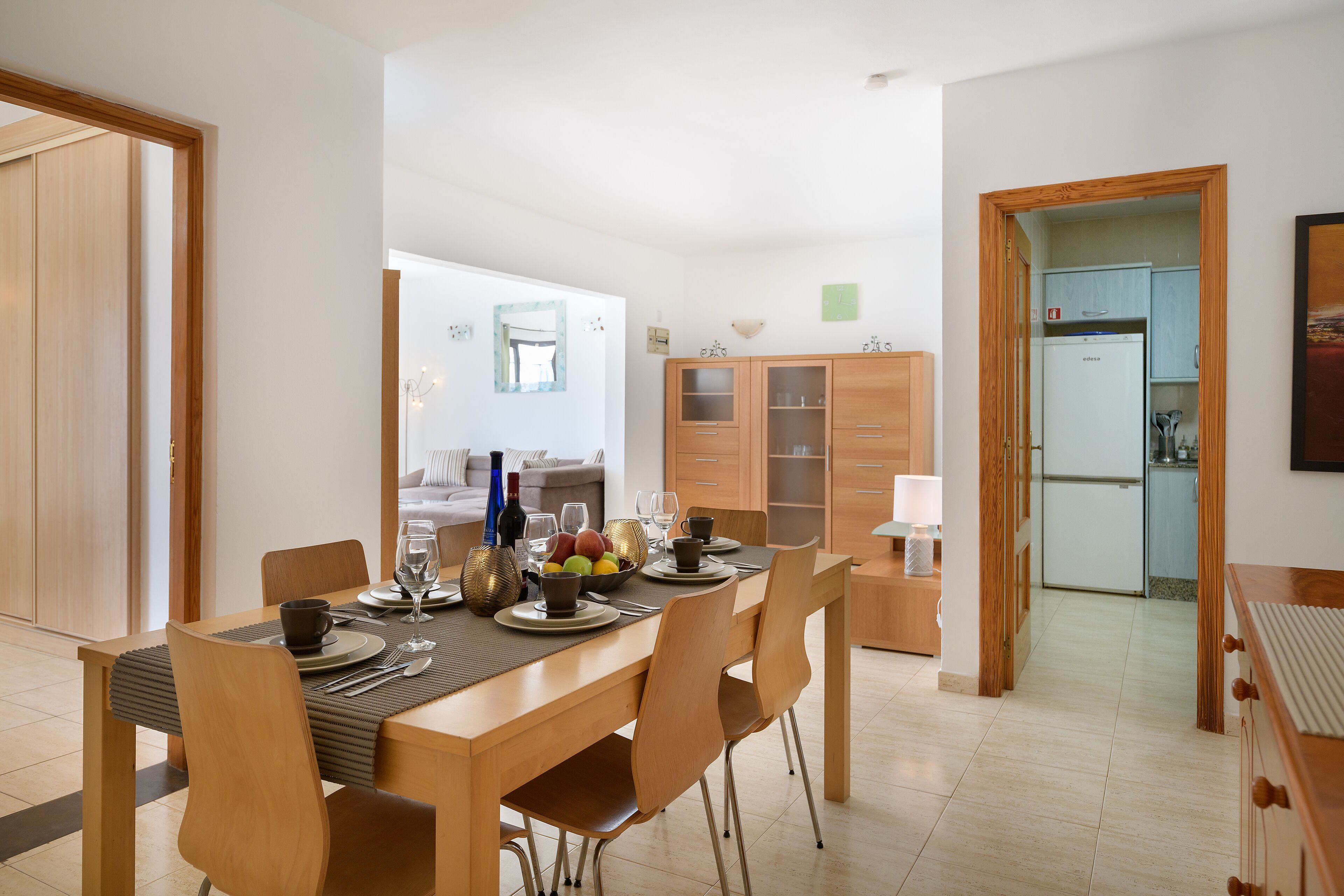 A dining area with a wooden table set for four, surrounded by chairs. There is a kitchen visible in the background with cabinets and a refrigerator.